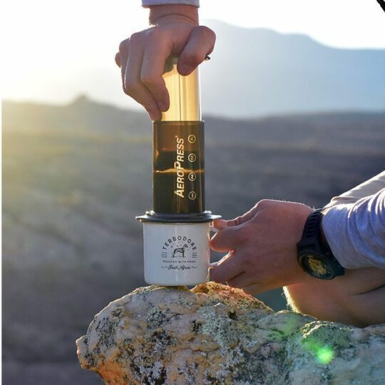 Man holding the Areopress and a Terbodore tin cup on a rock with mountains in the background