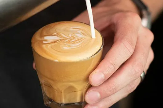 Man holding a glass mug with coffee inside, pouring milk into the mug - Terbodore